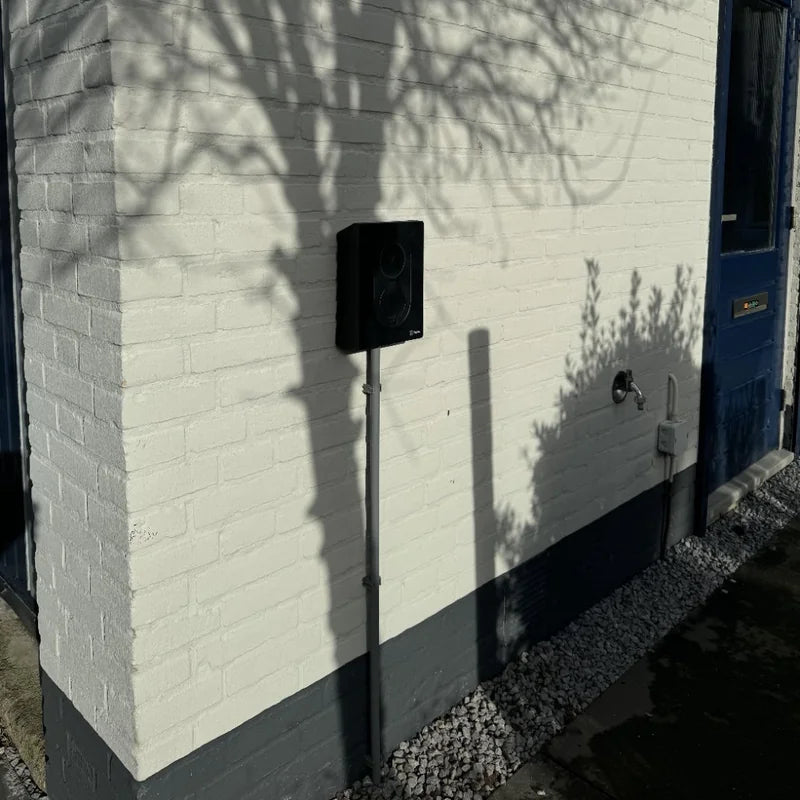Zwarte Zaptec Go laadpaal aan een wit geschilderde bakstenen gevel in de zon, met de schaduw van een boom erover.