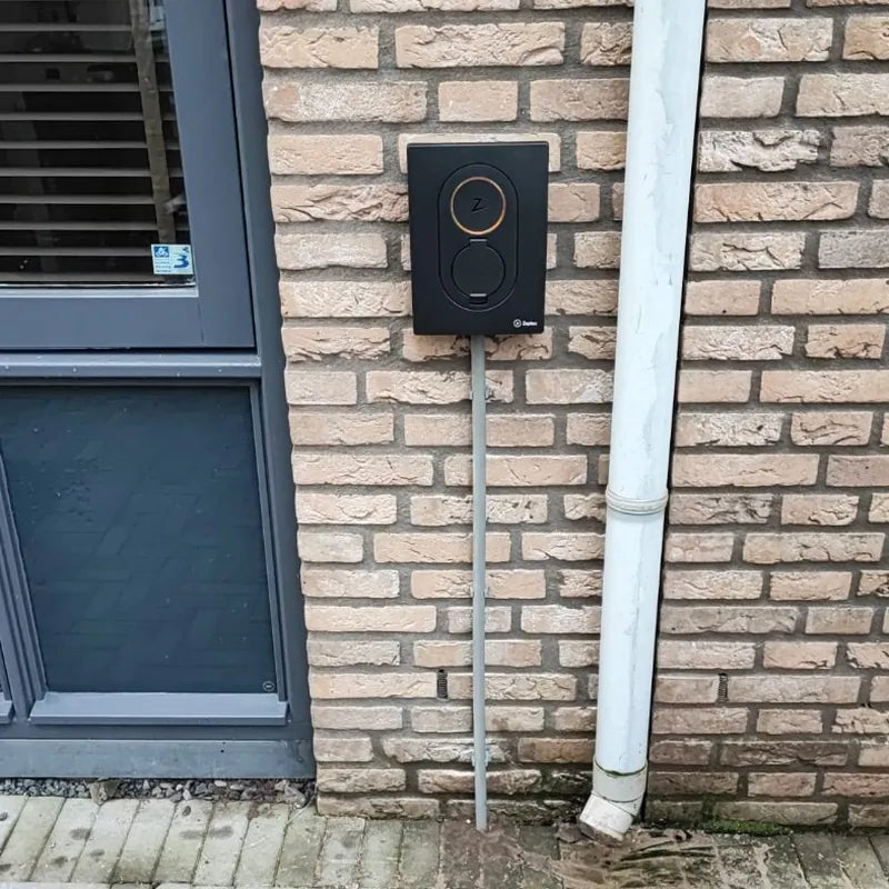 Zwarte Zaptec Go laadpaal aan een lichte bakstenen gevel naast de voordeur, met een grijze kabelbuis naar de grond.
