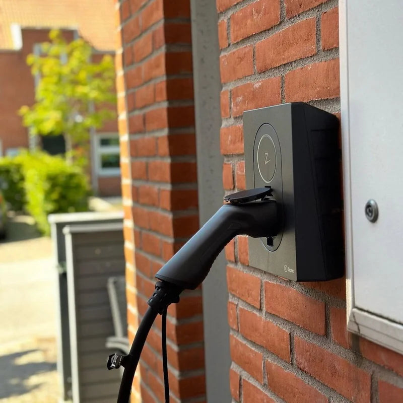 Zwarte Zaptec Go laadpaal aan een rode bakstenen muur, met een aangesloten Type 2-laadstekker in de contactdoos.